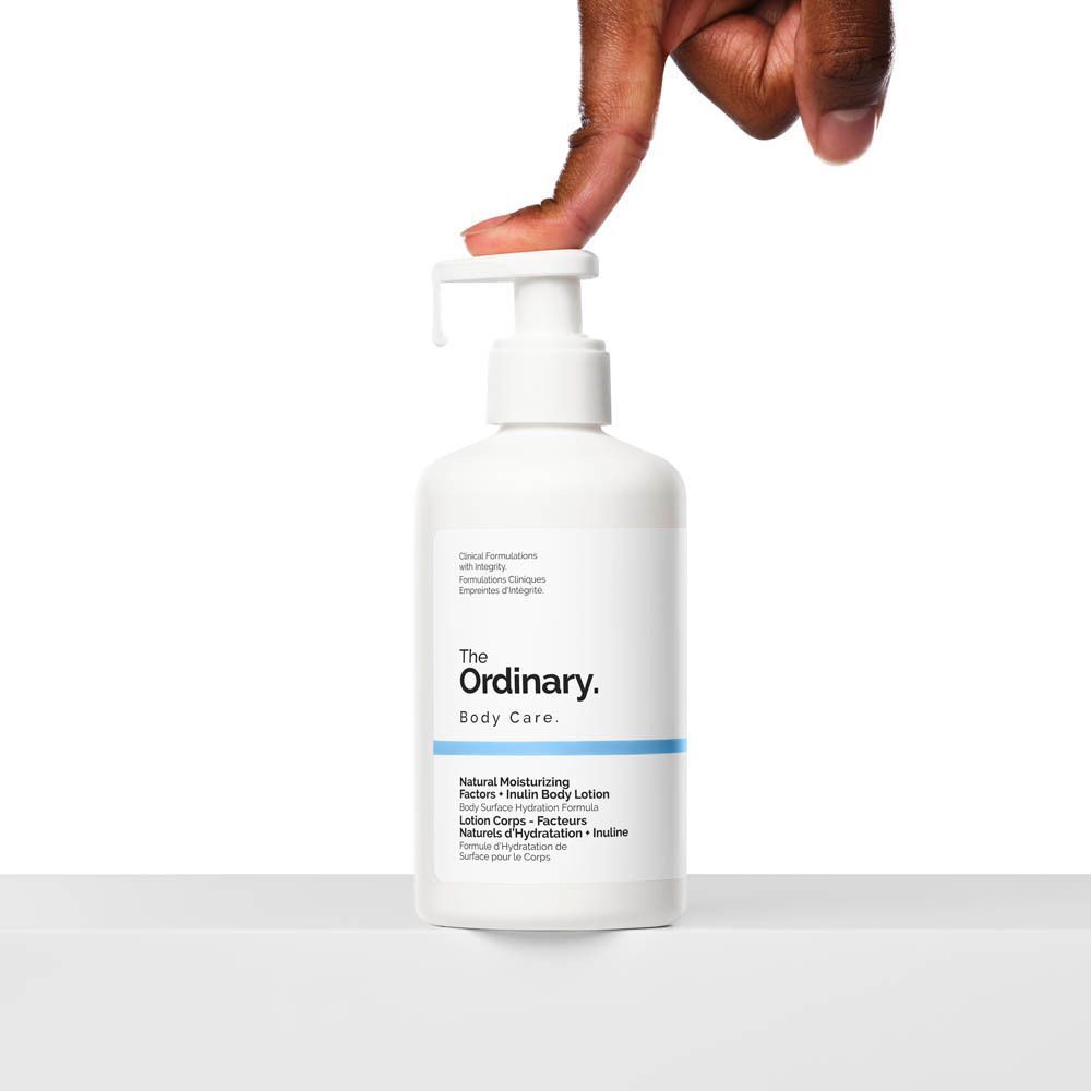 Weiße Flasche mit Pumpverschluss. Aufschrift: The Ordinary, Body Care, Natural Moisturizing Factors + Inulin Body Lotion. Hand drückt auf den Pumpkopf.