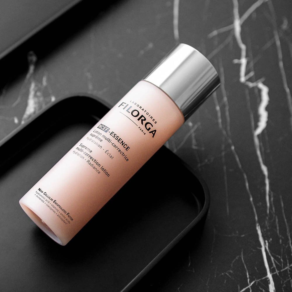 Un flacon avec bouchon argenté sur un plateau noir. Le flacon porte l'inscription "FILORGA" et "NCEF-ESSENCE Lotion multi-correctrice suprême".
