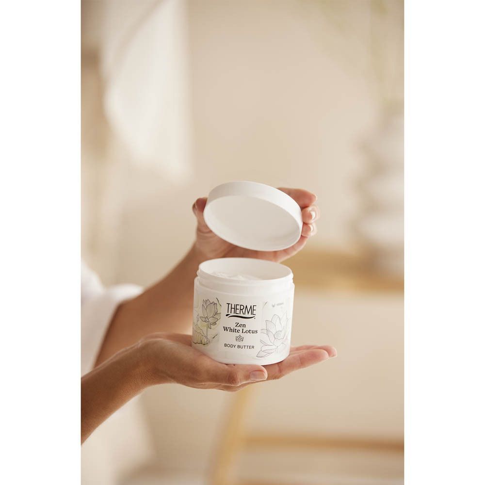 Mains tenant un pot blanc ouvert de Therme Zen White Lotus Body Butter. Couvercle à côté.