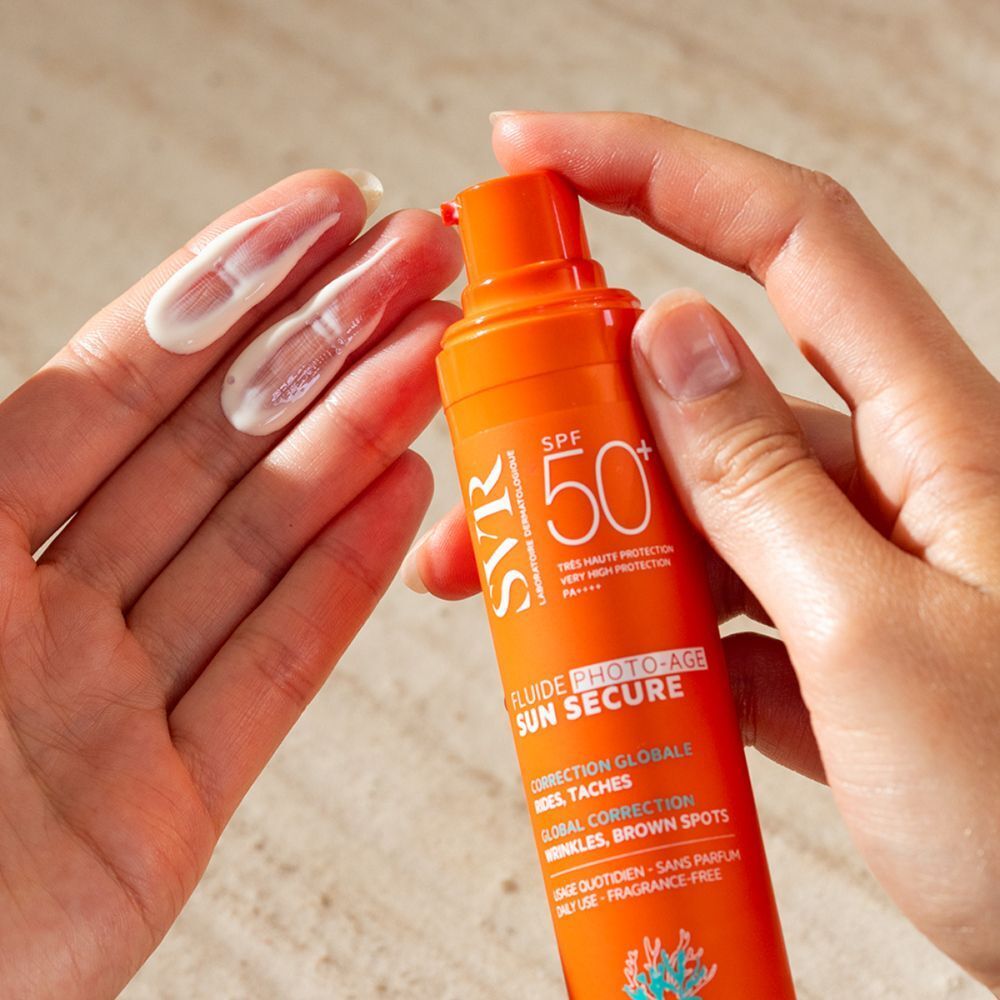 Handen houden een oranje fles SVR Sun Secure Fluide Photo-Age SPF50+ vast. Er zit crème op de handpalm.