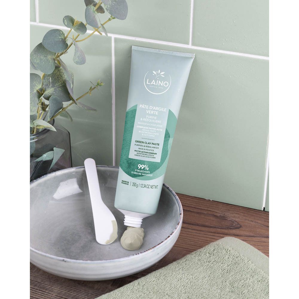Un tube de Laino Pâte Argile Verte, à côté d'un bol et d'une spatule. Produit placé sur un carrelage vert.