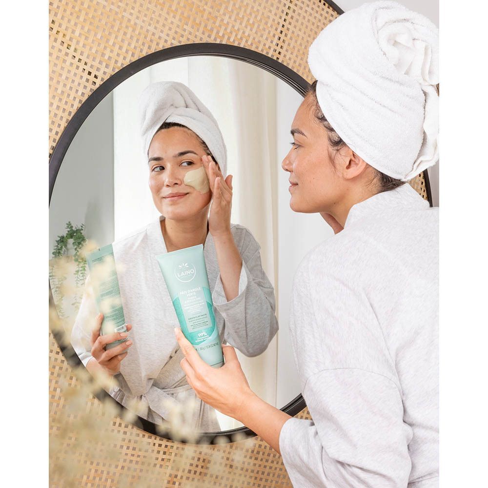 Femme appliquant Laino Pâte Argile Verte sur son visage. Elle se regarde dans le miroir. Un produit est visible dans sa main.