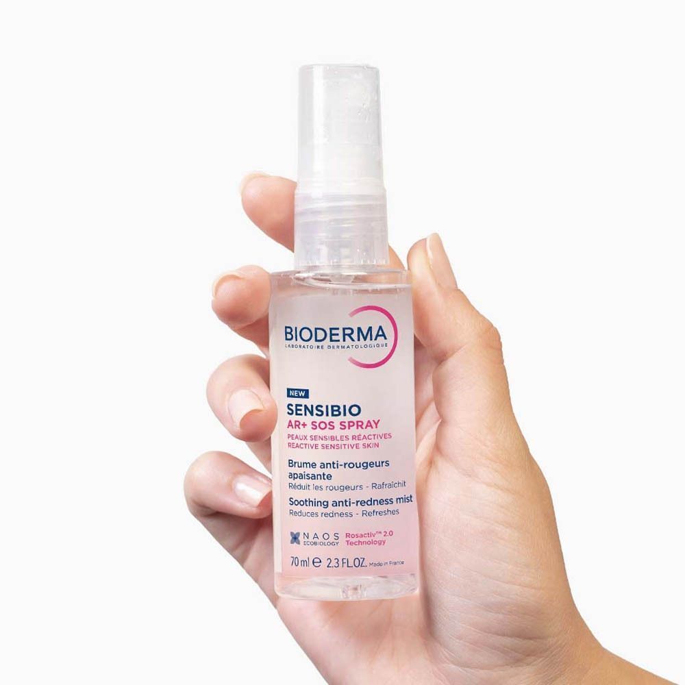 Hand houdt Bioderma Sensibio AR+ SOS Spray. Witte flacon met verstuiver, roze label, tekst.
