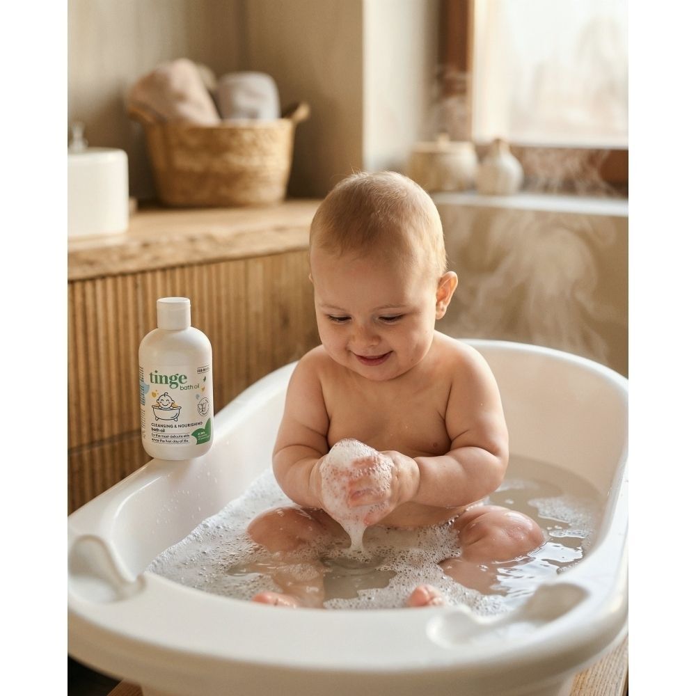 Un bébé assis dans une baignoire pour bébé avec de la mousse. Une bouteille de produit lavant pour bébé Tinge est à côté.