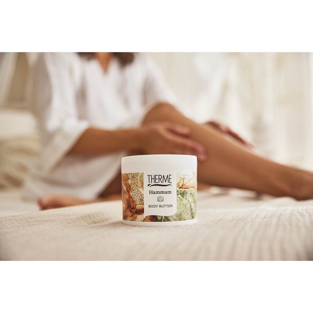 Un pot de Beurre Corporel Therme Hammam sur un lit, une femme se masse les jambes.
