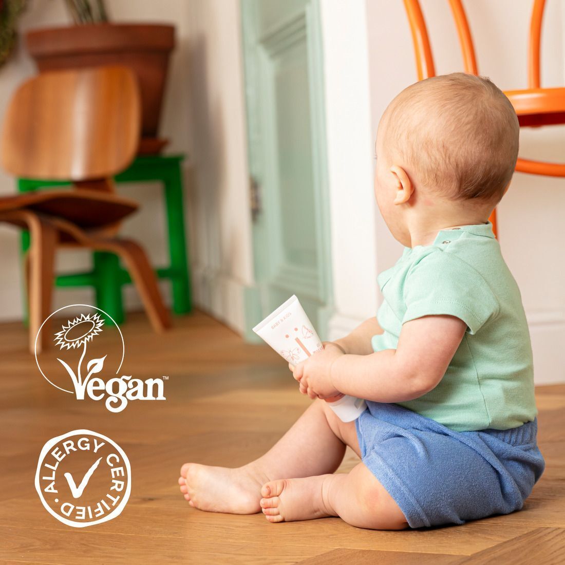 Ein Baby sitzt auf dem Boden und hält eine Flasche Naïf Baby Powder. Siegel für Vegan und Allergy Certified.