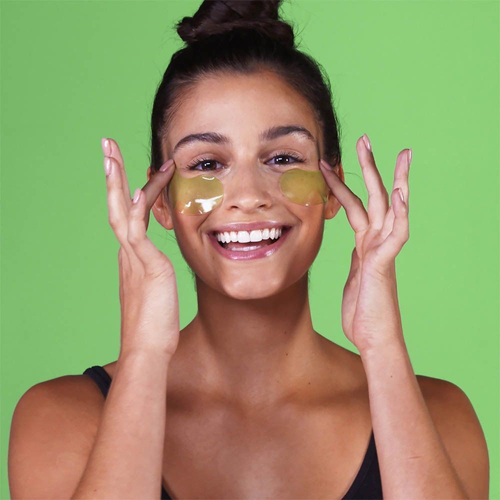Vrouw met groene oogpads onder haar ogen, lachend, tegen een groene achtergrond.