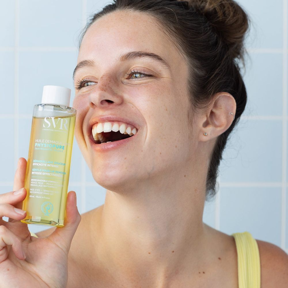 Een lachende vrouw houdt een fles SVR Physiopure Make-up Remover Olie vast.