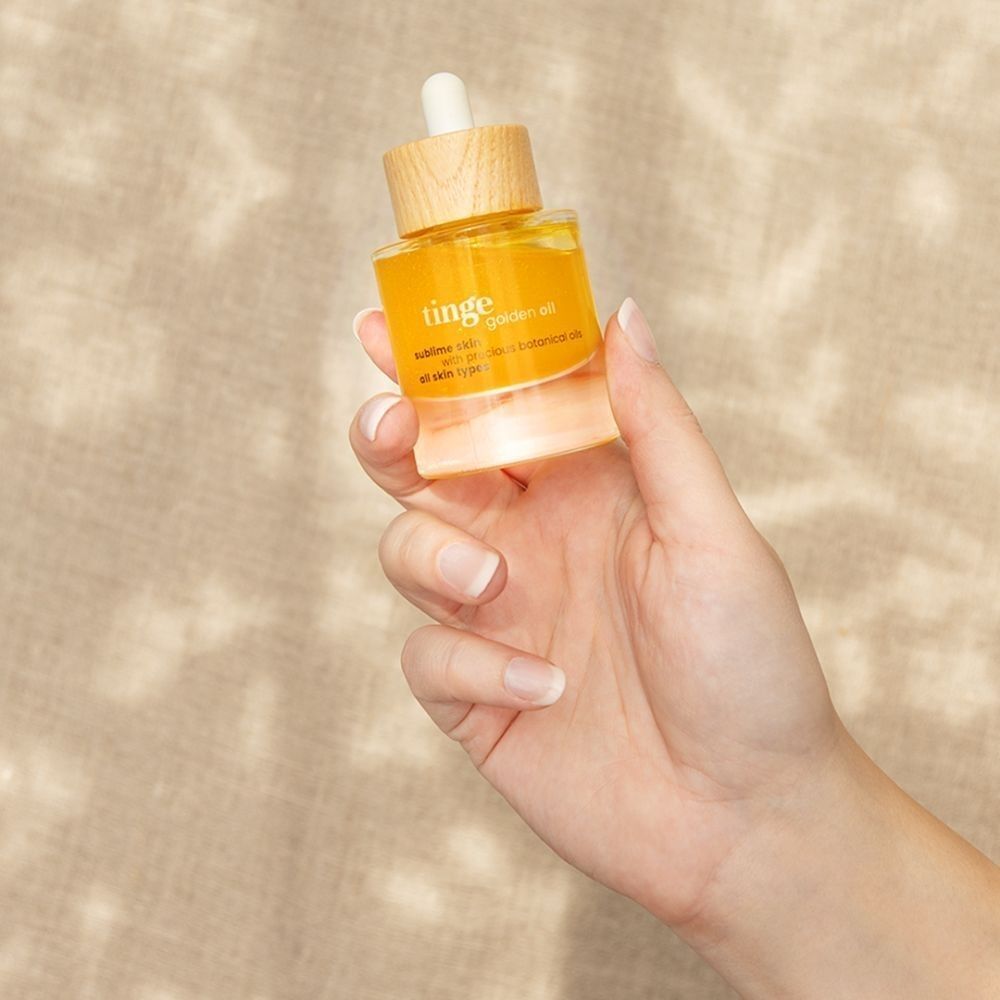 Een hand houdt een flesje Tinge Golden Oil met pipet en houten dop vast.