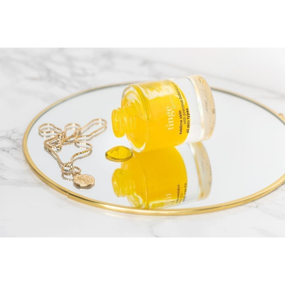 Een fles Tinge Golden Oil ligt op een spiegel, met een gouden ketting ernaast.