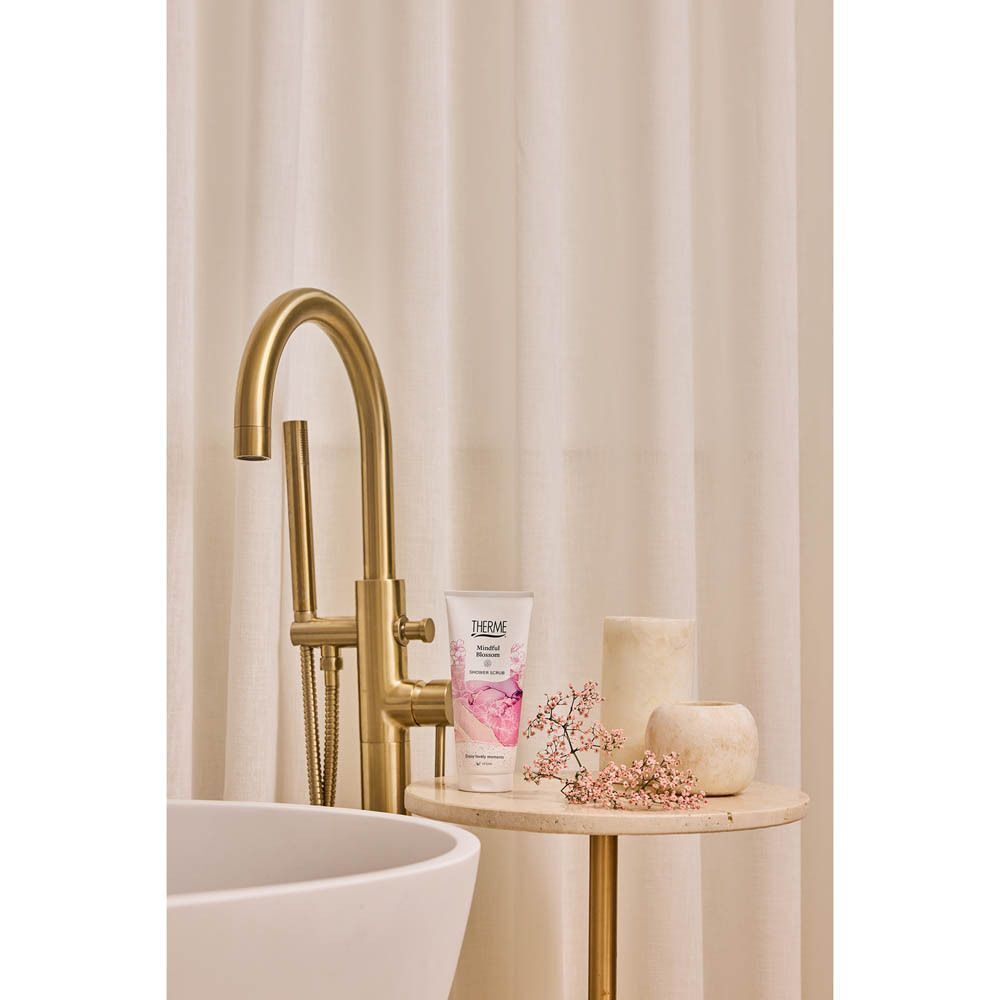 Een tube Therme Mindful Blossom Shower Scrub op een bijzettafel naast een badkuip.