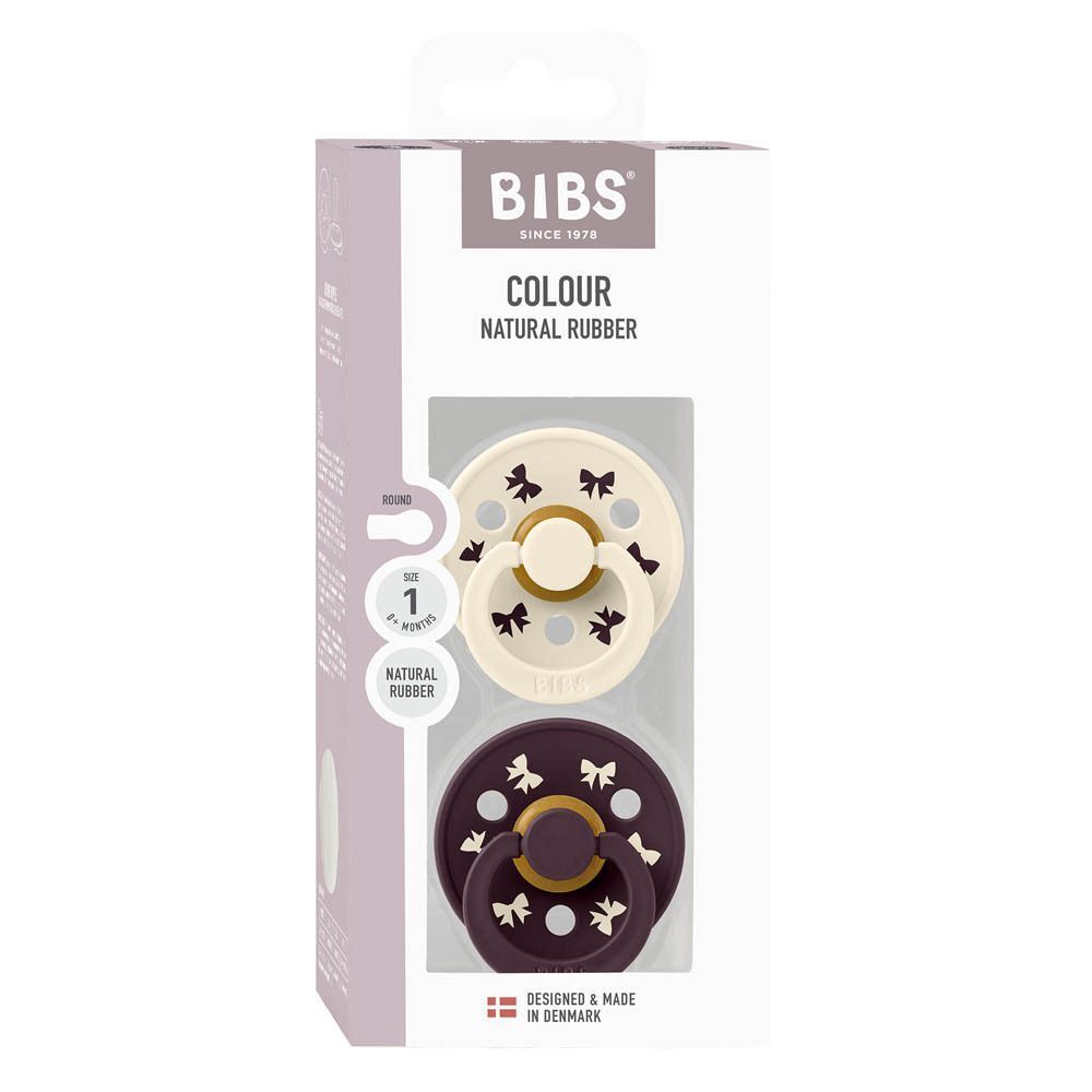 Deux sucettes BIBS Colour en caoutchouc naturel, une crème et une violet foncé avec motif nœud.