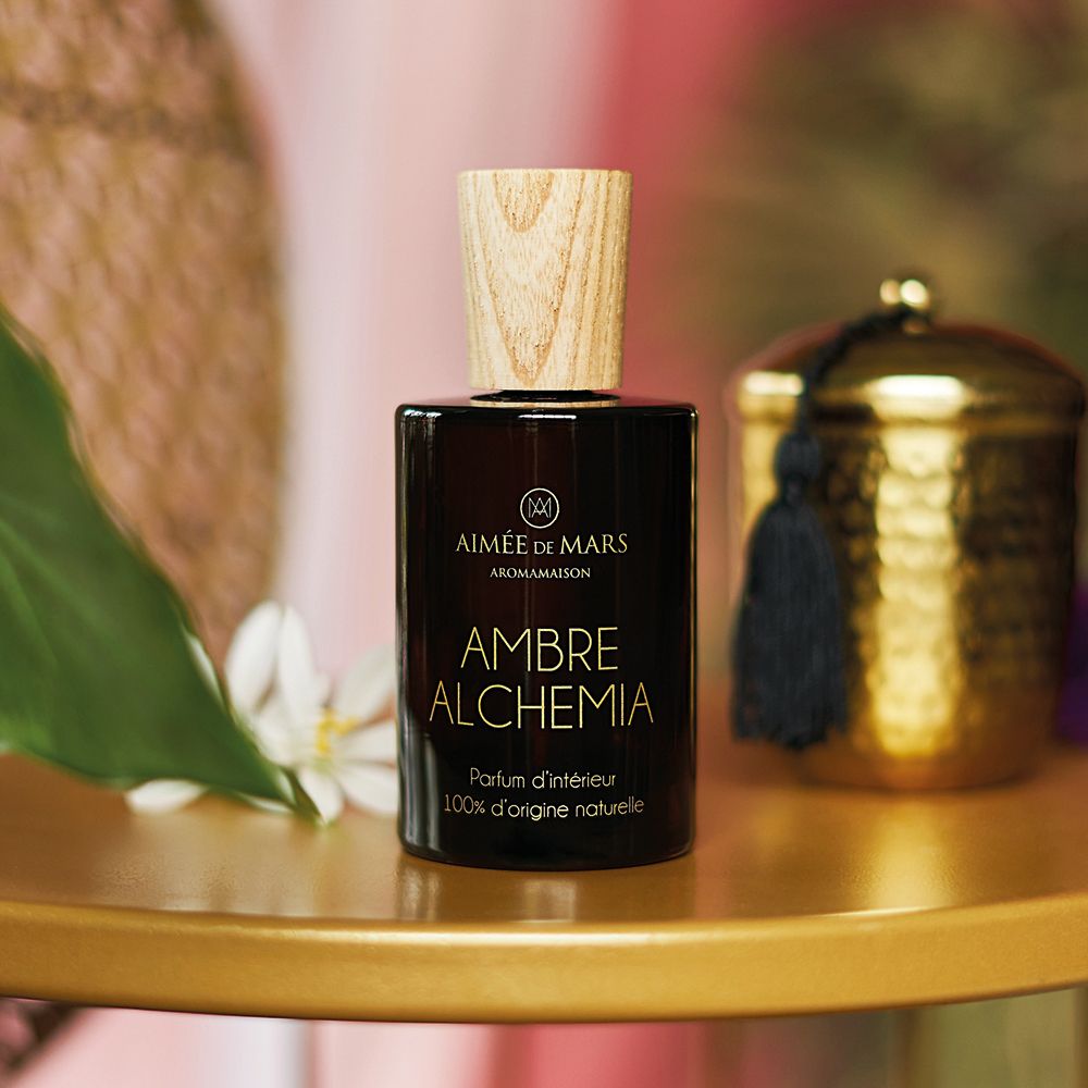 Braune Glasflasche mit Holzverschluss. Aufschrift: AMBRE ALCHEMIA, Parfum d'intérieur, 100% d'origine naturelle. Auf goldenem Tisch.