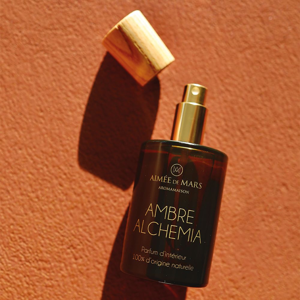 Braune Glasflasche mit Holzverschluss, Deckel daneben. Aufschrift: AMBRE ALCHEMIA, Parfum d'intérieur, 100% d'origine naturelle.