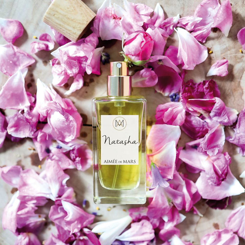Aimée de Mars Natasha Eau de Parfum Flakon auf Holz. Umgeben von rosa Blütenblättern. Goldener Sprühkopf.