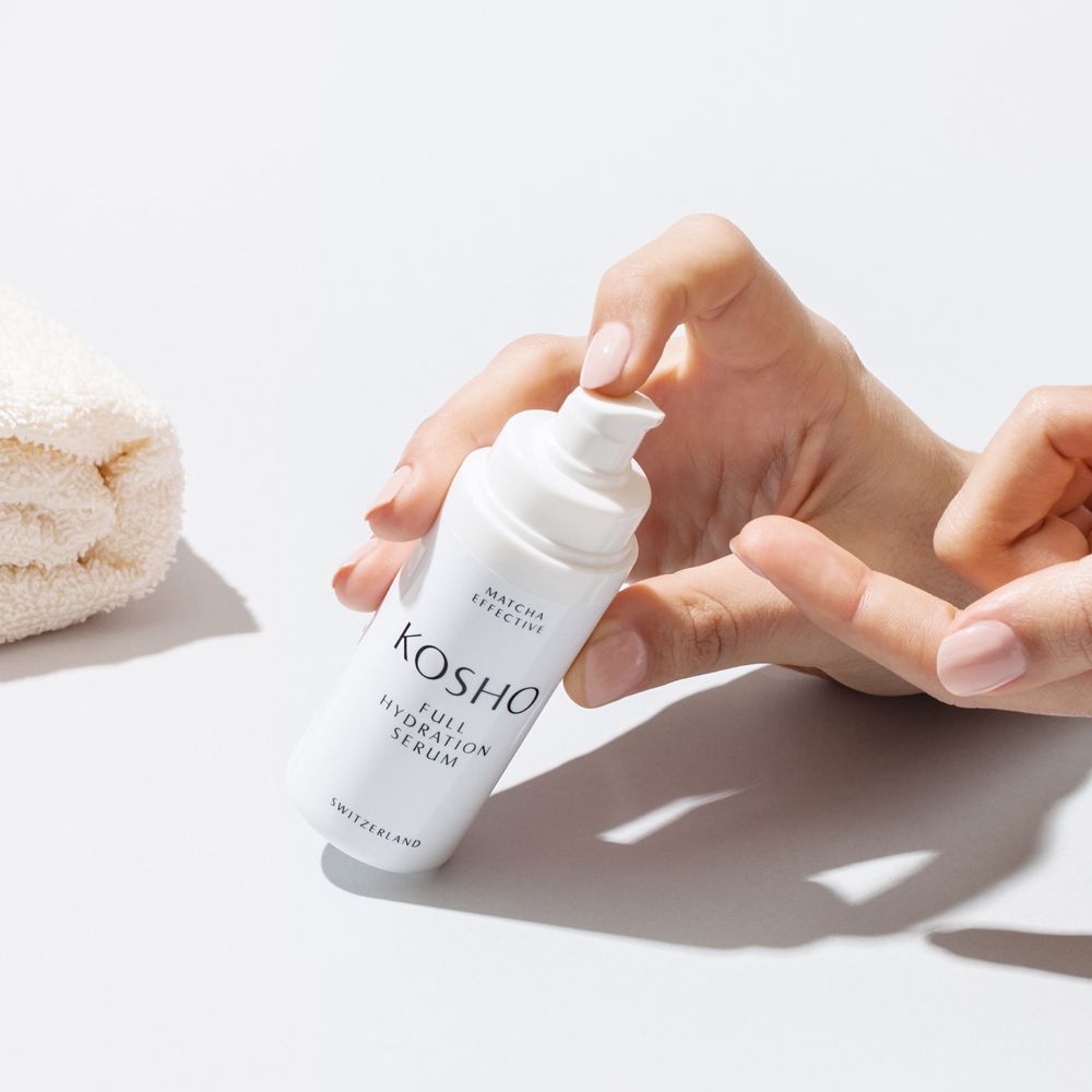 Flacon de sérum blanc avec inscription KOSHO Full Hydration Serum. Mains tenant le flacon et une serviette.