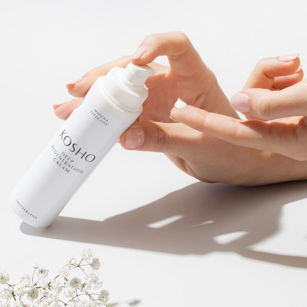Hand hält weiße Flasche KOSHO Deep Regeneration Cream. Creme wird auf Finger aufgetragen. Weiße Blüten.