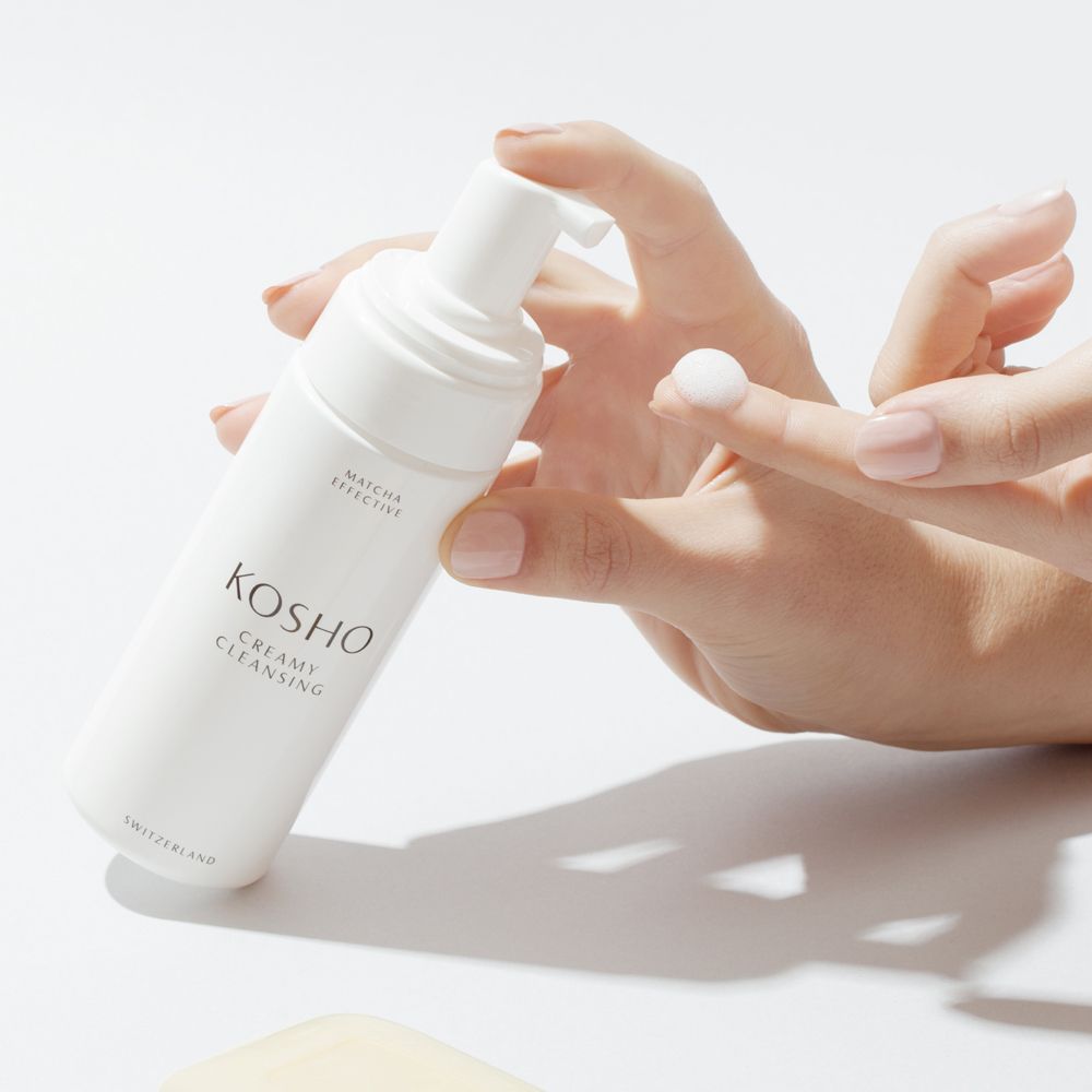 Mains tenant un flacon blanc KOSHO Creamy Cleansing. Une noisette de mousse sur le doigt. Produit sur fond clair.