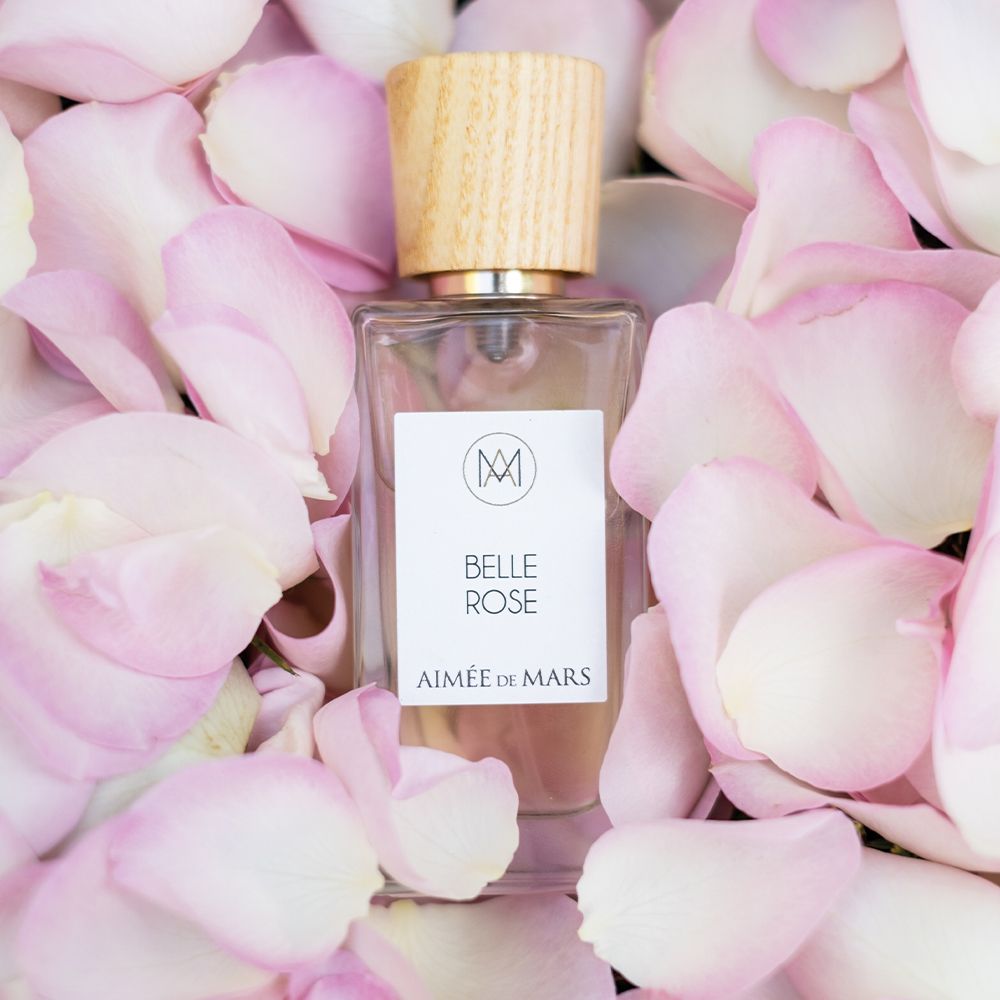 Aimée de Mars Belle Rose Eau de Parfum Flakon, umgeben von rosa Rosenblättern. Rechteckige Glasflasche mit Holzverschluss.