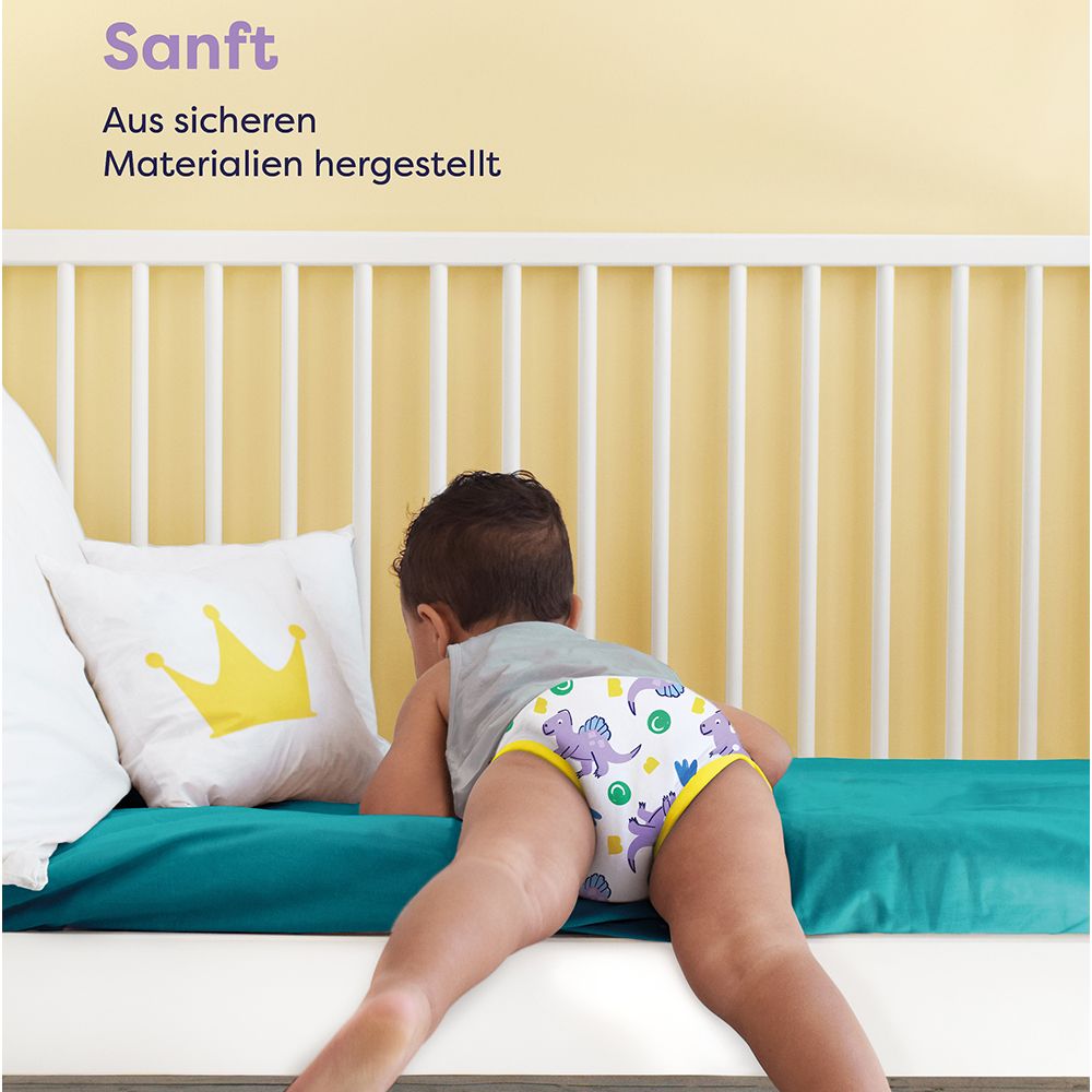Enfant avec culotte d'apprentissage sur le lit. Motif : dinosaure. Oreiller jaune avec couronne.