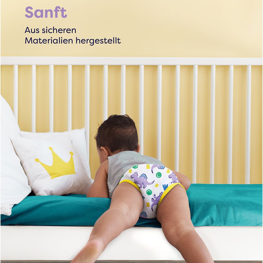Kind mit Lernhöschen auf Bett. Text: Sanft. Aus sicheren Materialien hergestellt. Gelbes Kissen mit Krone.