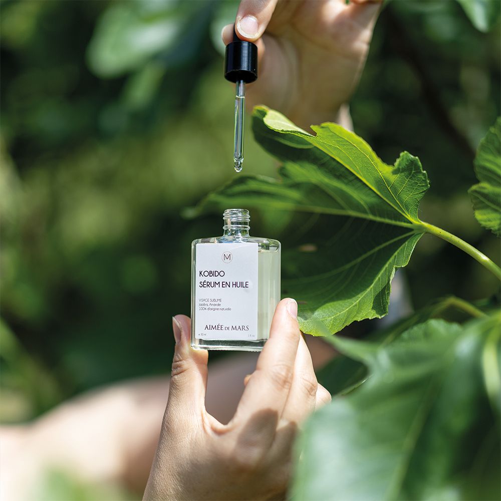 Hände halten eine Glasflasche mit Ölserum und Tropfer über grünen Blättern. Tropfen fallen aus dem Tropfer.