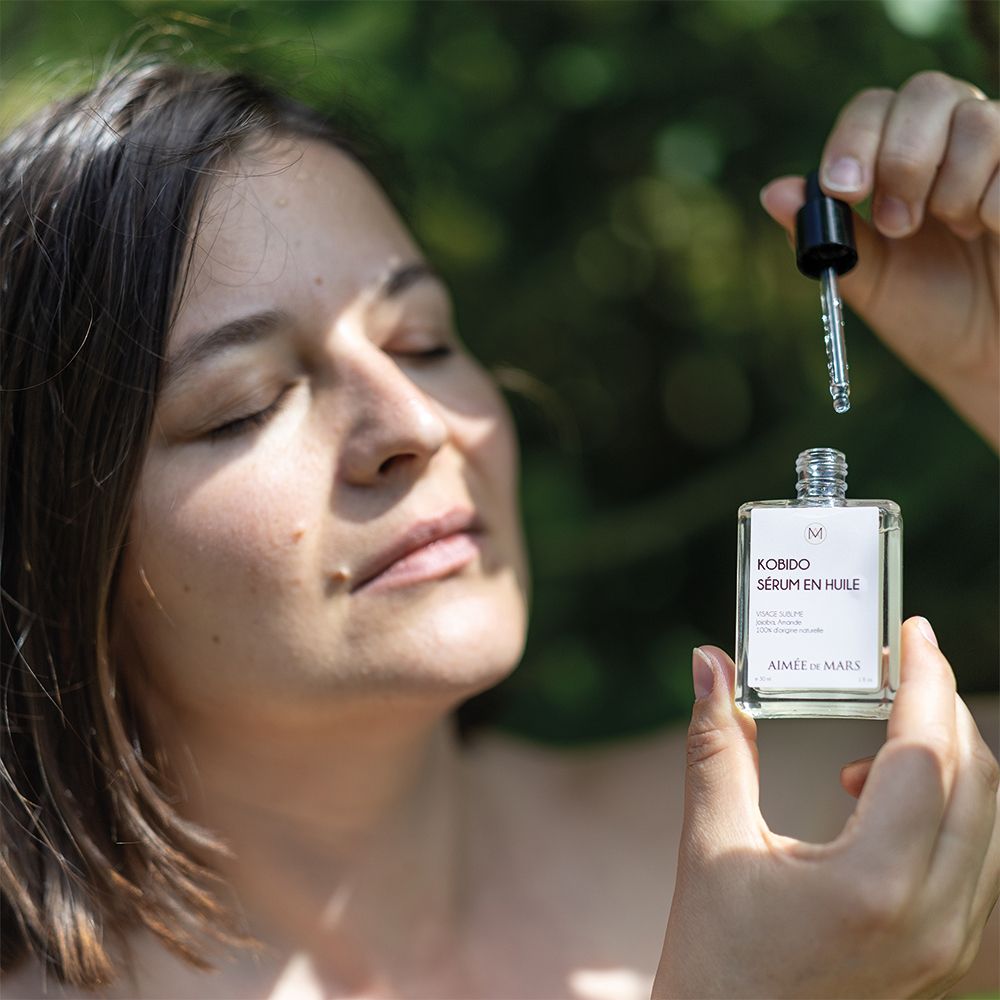 Frau hält eine Glasflasche mit Ölserum und Tropfer. Gesicht entspannt, Augen geschlossen. Grüner Hintergrund.