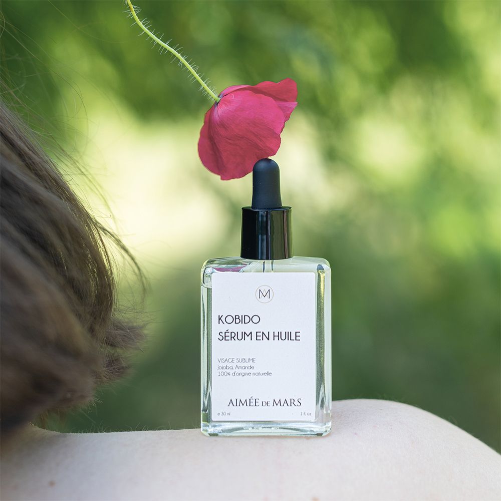 Glasflasche mit Ölserum und Tropfer, platziert neben einer roten Blume. Schulter einer Person im Hintergrund.