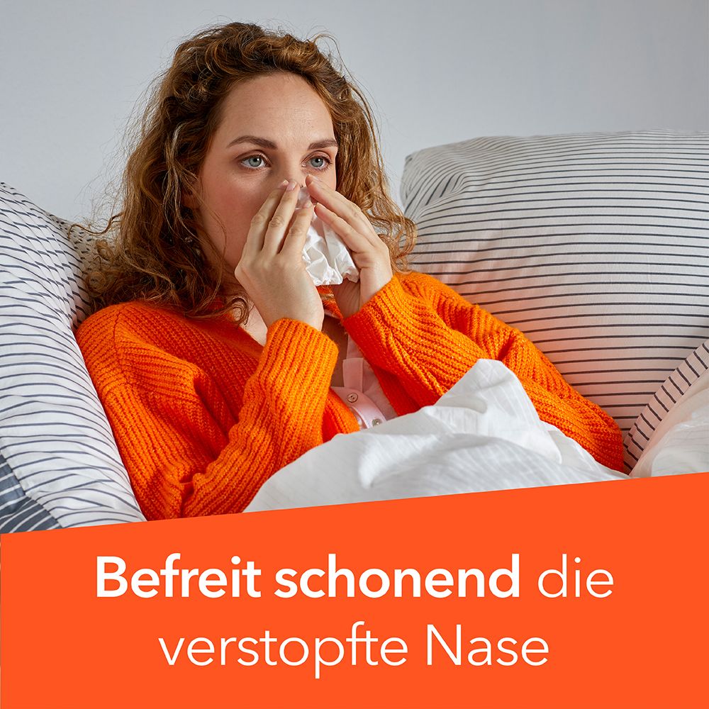 Frau mit orangefarbenem Pullover liegt im Bett und hält ein Taschentuch an die Nase. Befreit schonend die verstopfte Nase.
