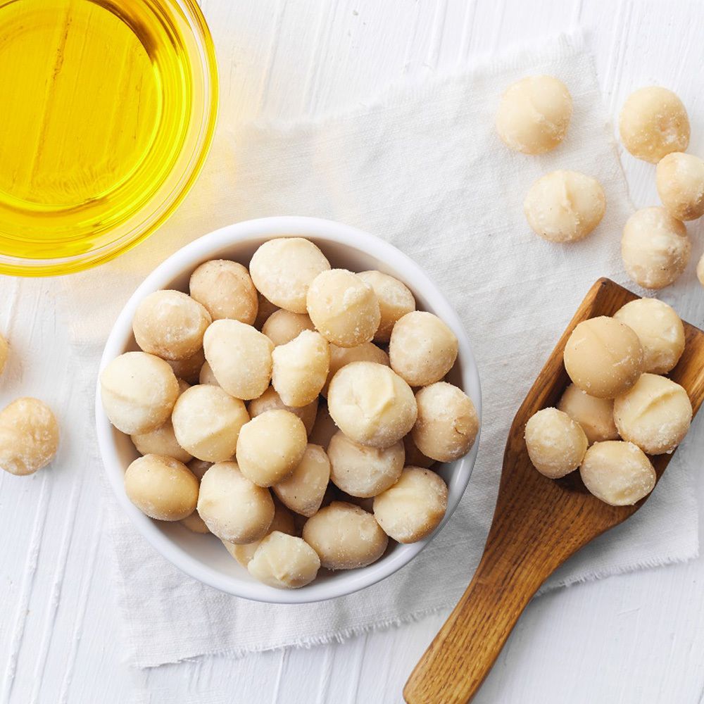 Bol blanc avec noix de macadamia, cuillère en bois et huile à côté.