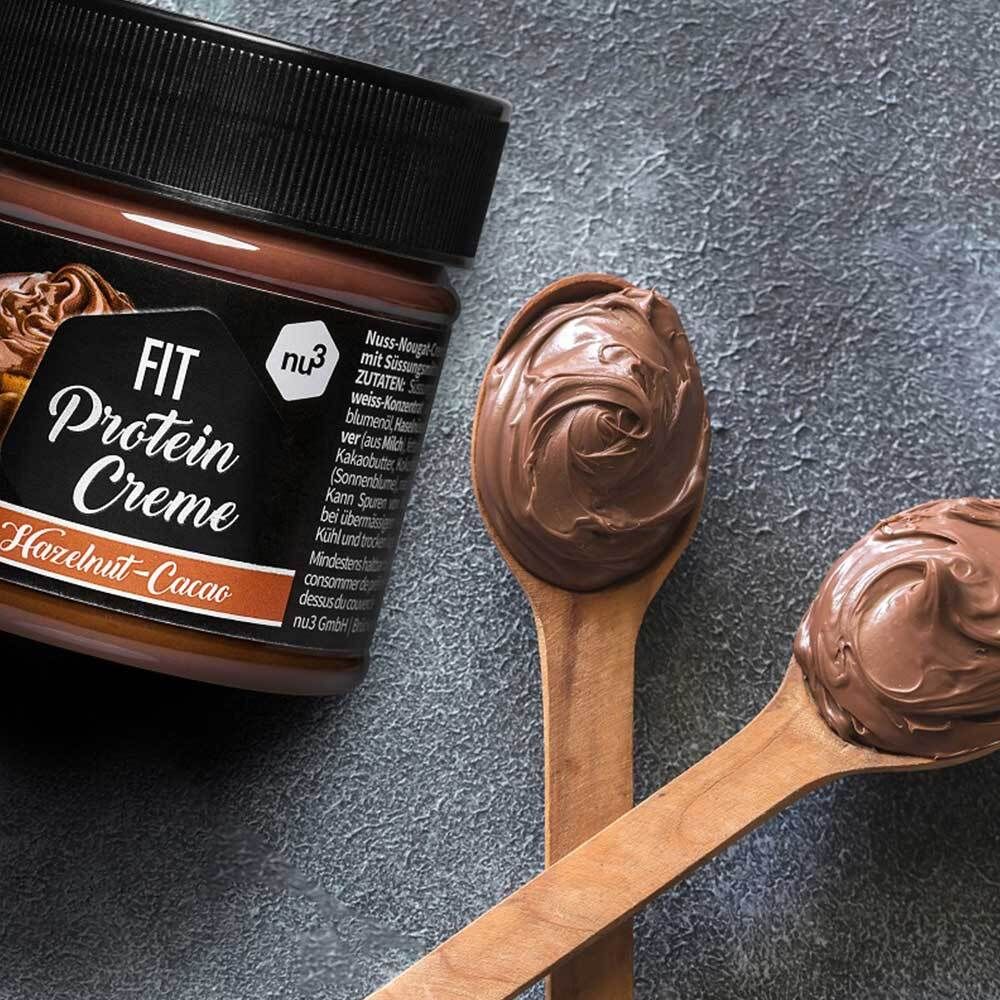 Deux cuillères en bois avec crème protéinée Fit. Pot de crème en arrière-plan.