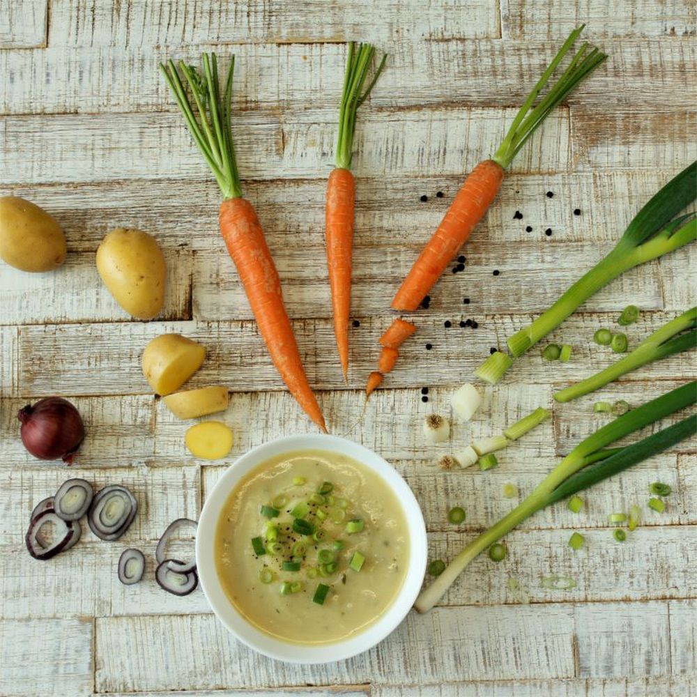 Vue de dessus des ingrédients de la soupe de pommes de terre : carottes, oignons, poireaux, pommes de terre et un bol de soupe.