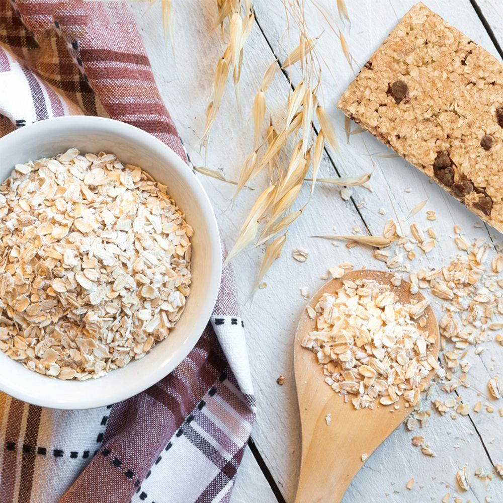 Energieriegel neben einer Schüssel mit Haferflocken, einem Holzlöffel und Ähren auf weißem Holz. Textur.