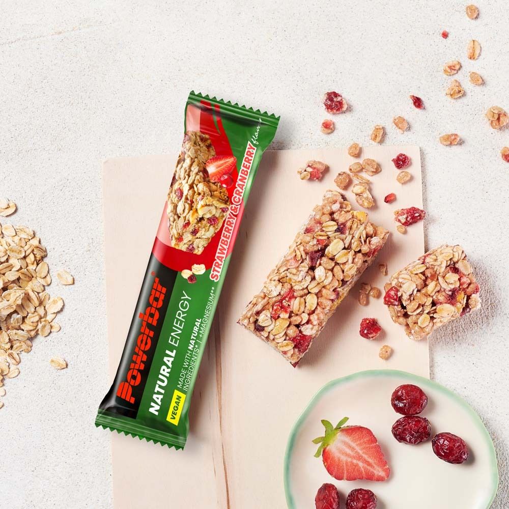 PowerBar Natural Energy Riegel, Erdbeer-Cranberry. Auf hellem Hintergrund. Riegel und Zutaten verstreut. Vegan. Riegel in Verpackung.