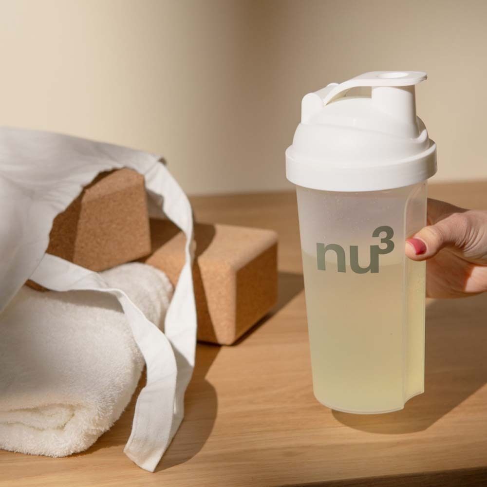 Eine Person hält einen weißen Shaker mit nu3-Logo. Auf einem Holztisch liegen Yogablöcke, eine Tasche und ein Handtuch.