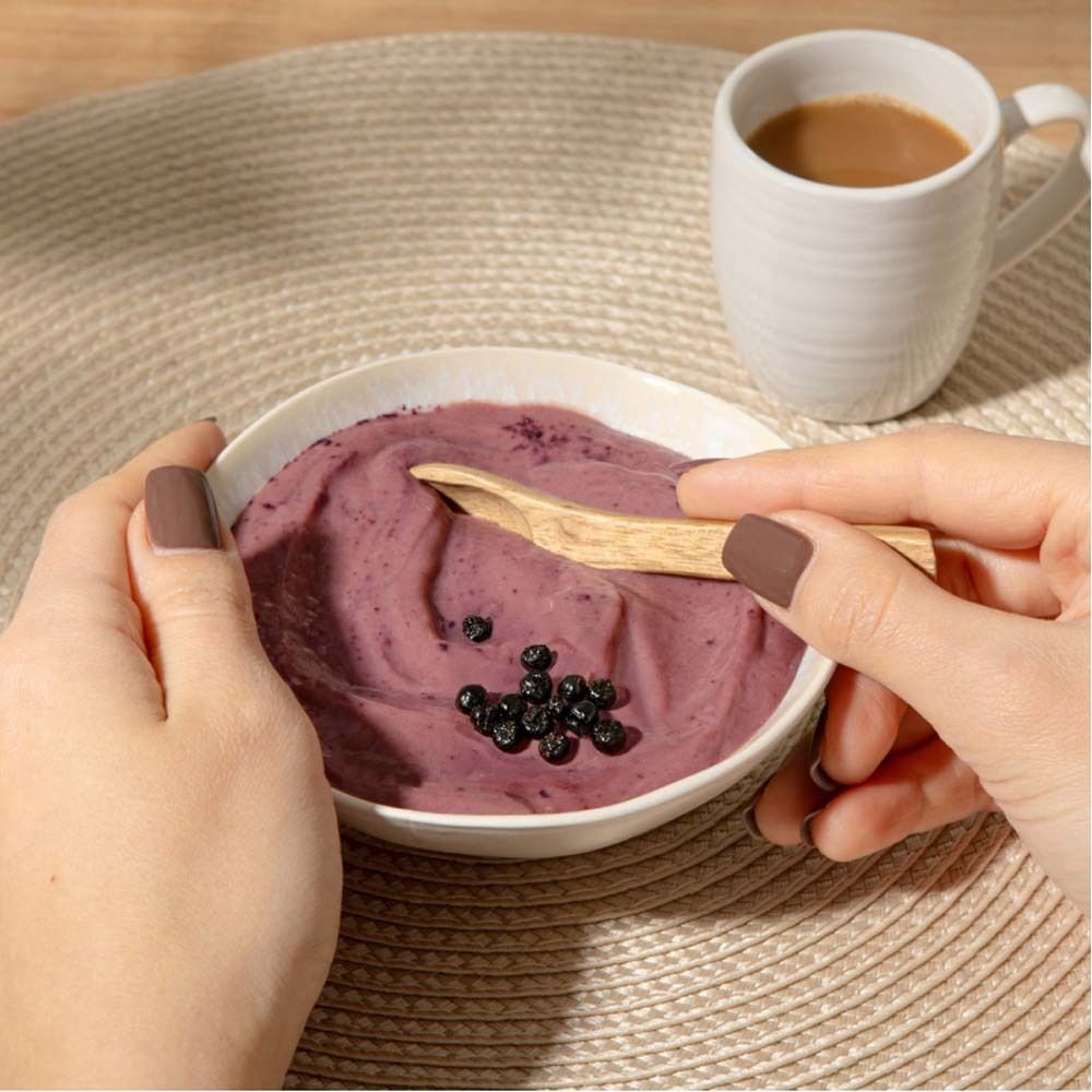 Nahaufnahme einer Schüssel mit lila Smoothie, Aroniabeeren und einem Holzlöffel. Eine Tasse Kaffee steht daneben. Hände halten die Schüssel.