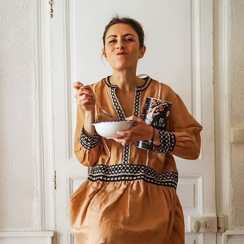 Frau mit einem Löffel und einer Dose nu3 Fit Müsli. Sie sitzt vor einer weißen Tür und isst Müsli aus einer Schüssel.