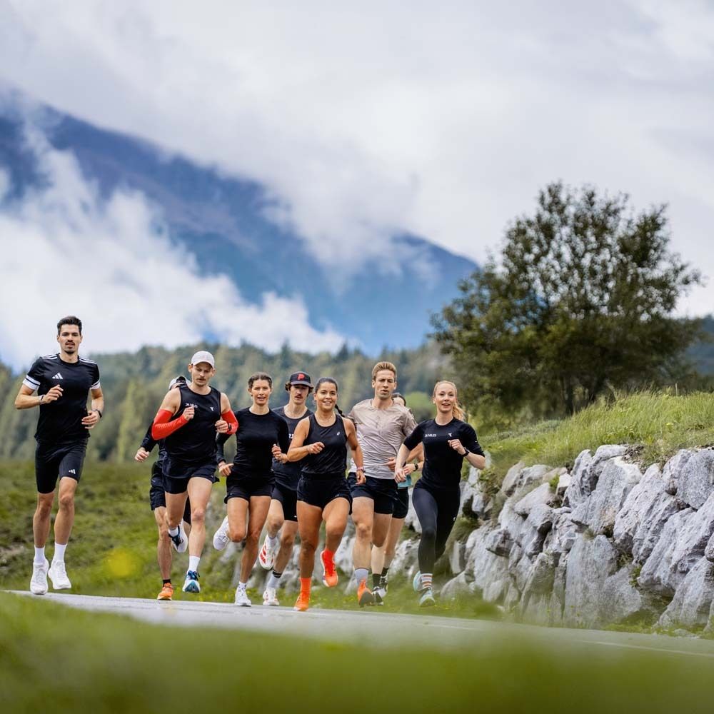 Gruppe von Läufern auf einer Straße in den Bergen. Sie tragen Sportkleidung. Im Hintergrund Berge und Bäume.