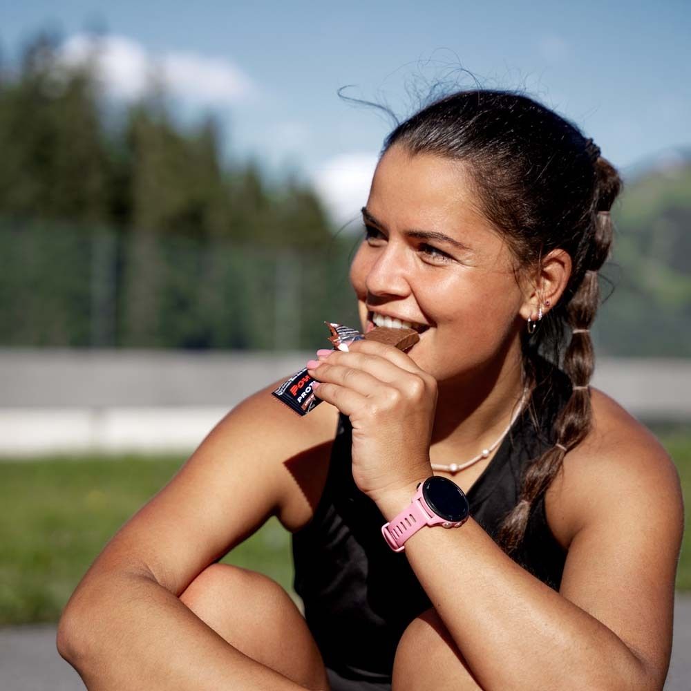 Frau isst Proteinriegel. Riegel in schwarzer Verpackung. Sie trägt eine Smartwatch.