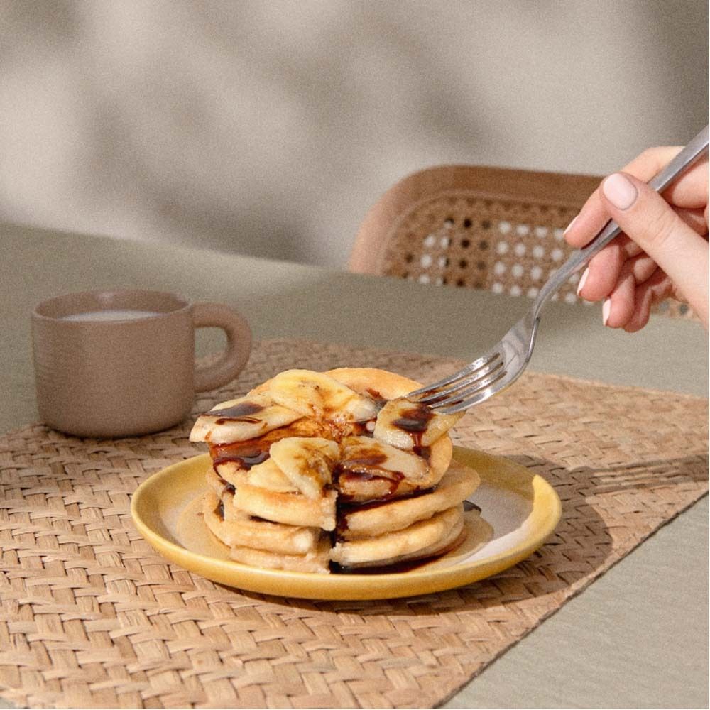 Pfannkuchen auf gelbem Teller, mit Gabel. Aufguss mit Schokosauce und Bananenscheiben. Hintergrund: Kaffeetasse.