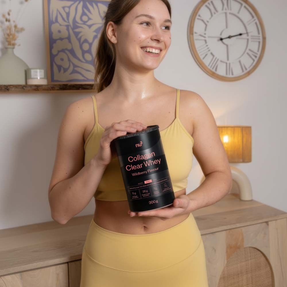 Frau hält eine schwarze Dose nu3 Collagen Clear Whey. Gelbes Sport-Outfit. Hintergrund: Zimmer mit Uhr und Möbeln.