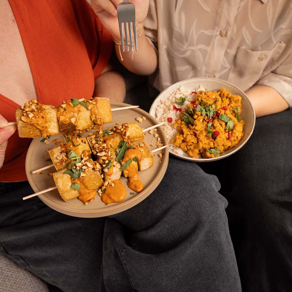 Personen essen Tofu-Spieße mit Erdnuss-Saté-Sauce und Linsen-Curry. Auf einem Teller sind Spieße, in einer Schüssel Curry.