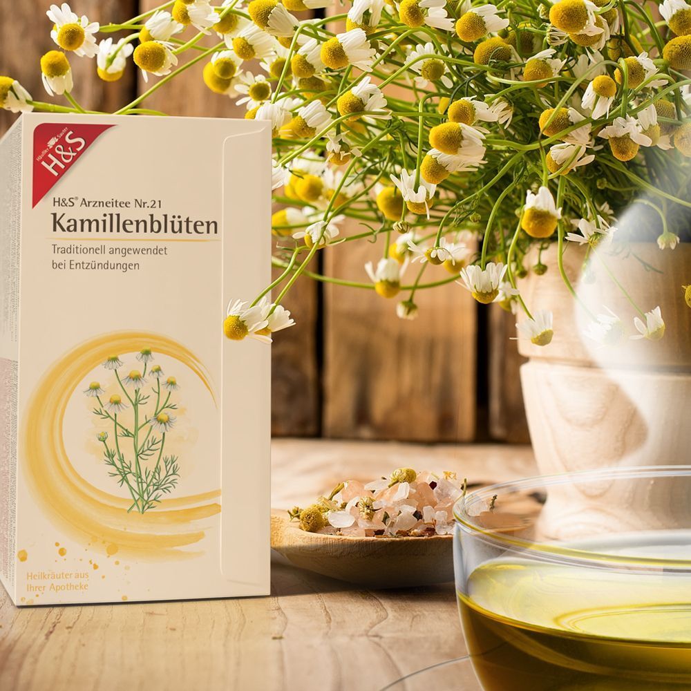 H&S Kamillenblüten Nr. 21 Packung neben Kamillenblüten und einer Tasse Tee. Weißer Karton mit rotem Logo und gelben Verzierungen. Text: Kamillenblüten, traditionell.