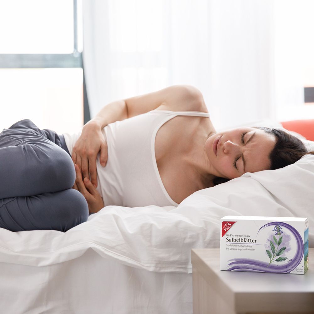 Frau liegt im Bett, hält sich den Bauch. H&S Salbeiblätter Packung liegt daneben.