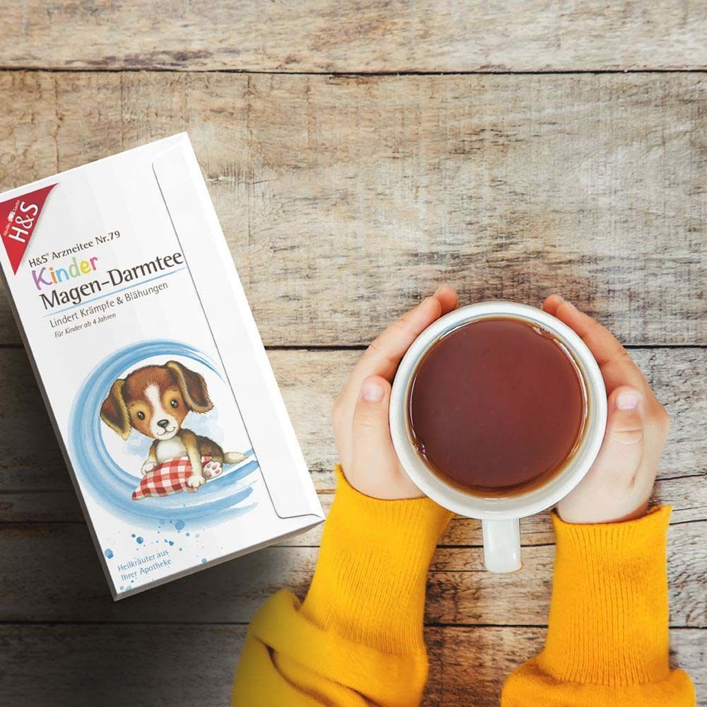 H&S® Kinder Magen-Darmtee Packung neben einer Tasse Tee. Hände halten die Tasse. Holzuntergrund.