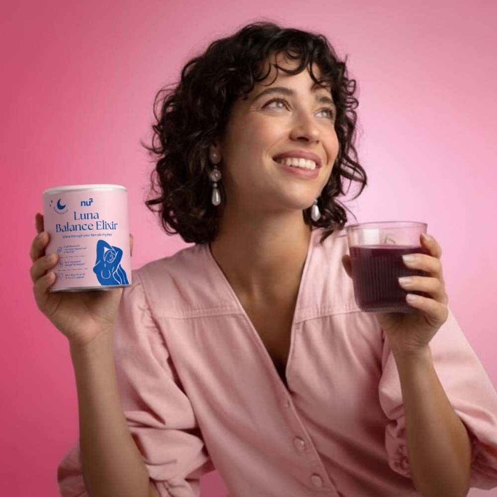 Frau hält eine Dose nu3 Luna Balance Elixir und ein Glas Getränk. Rosa Hintergrund. Die Frau lächelt und schaut nach oben.