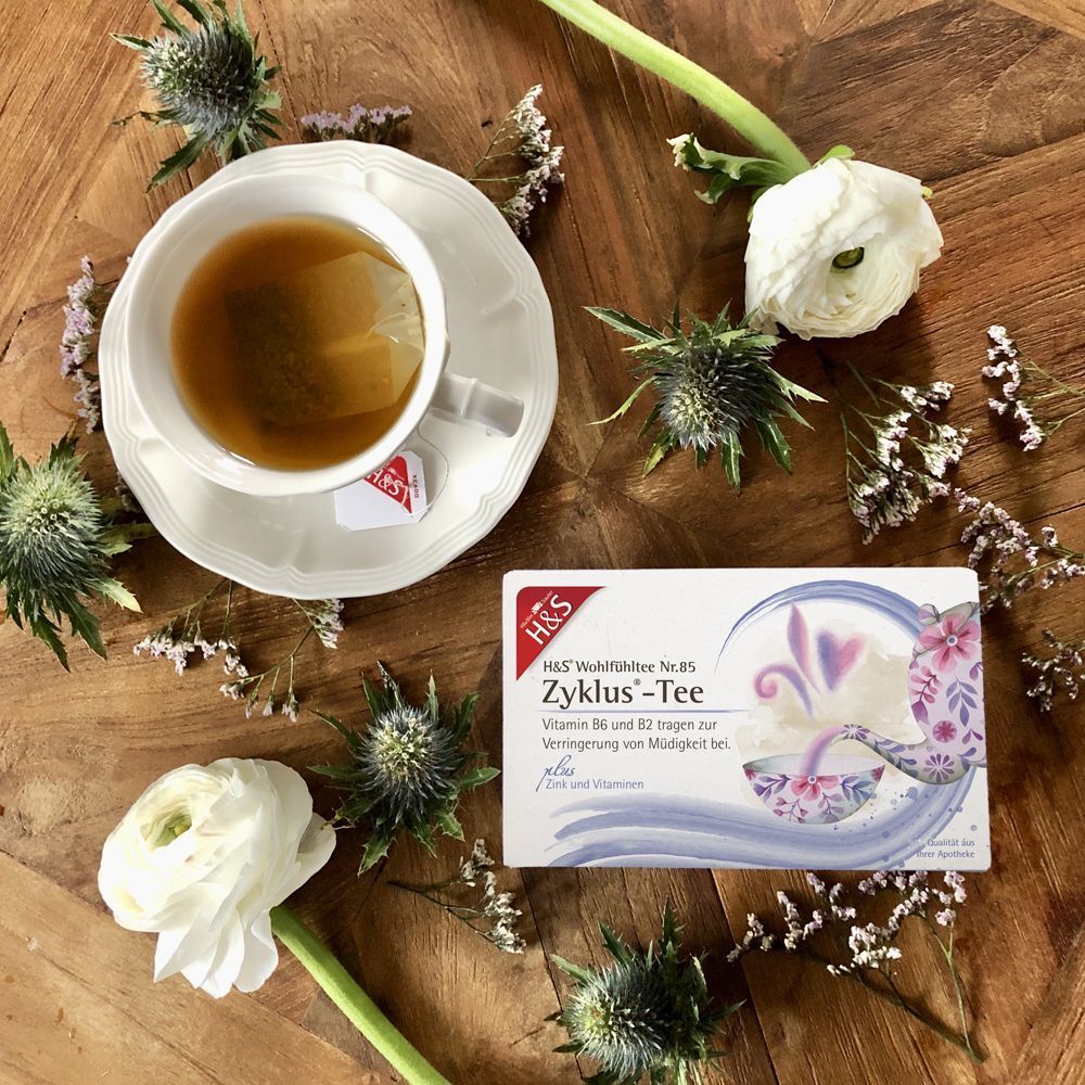 Eine Tasse Tee und eine Packung H&S Zyklus-Tee auf Holzoberfläche. Teebeutel im Tee. Dekoration mit Blumen.