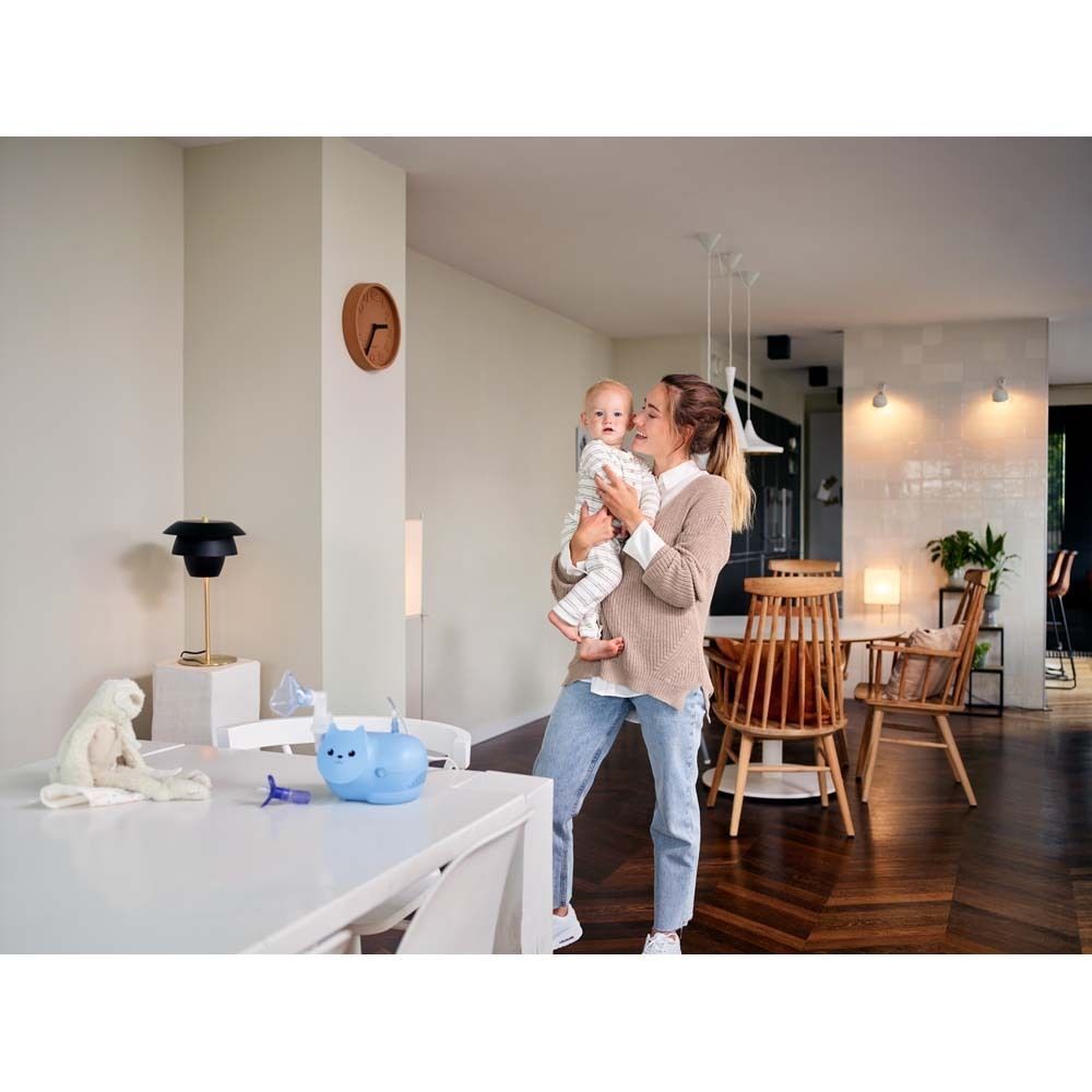 Frau hält Baby. Auf Tisch: OMRON Nami Cat Inhalationsgerät. Wohnzimmer-Szene.