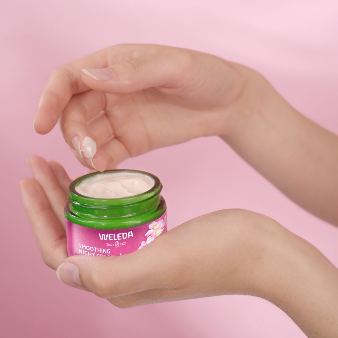 Hände halten einen Tiegel WELEDA Nachtpflege. Creme wird entnommen. Rosa Hintergrund.