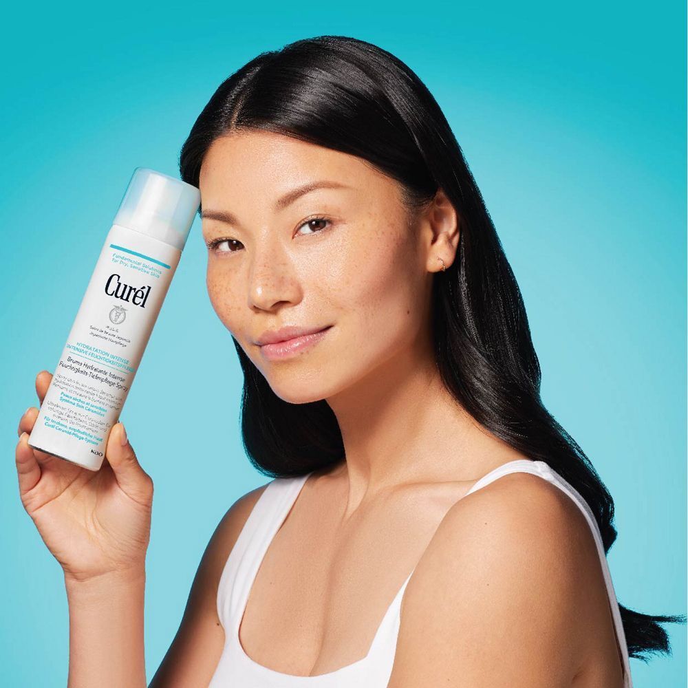 Asiatische Frau hält eine weiße Sprühflasche mit hellblauem Deckel. Aufschrift: Curel, Hydratation Intense, Feuchtigkeits-Tiefenpflege Spray.